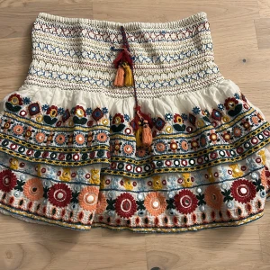 Broderad kortkjol med volang och tofsar - Superfin kortkjol i bomull med broderade blommor i rött, orange, blått och gult. Säljer då den inte kommer tillanvänding längre å har använt den ca 3 gånger