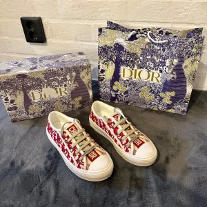 Dior sneakers med röd monogram - Snygga Dior sneakers med röd och vit monogramdesign, tydlig Christian Dior-logga på plösen och sidan. Skorna har vita snören, platt sula och rund tå. Tillverkade i canvas med detaljer i skinn och gummisula. Perfekta för dig som vill sticka ut med lyxig streetstyle.