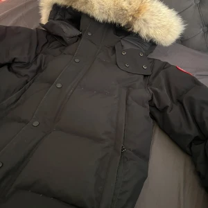 canada goose wyndham - Säljer en svart dunjacka från Canada Goose med stor huva och äkta pälsdetalj. Jackan har klassisk patch på ärmen, dragkedja och knappar framtill samt flera fickor. Perfekt för kalla vinterdagar och riktigt snygg passform.