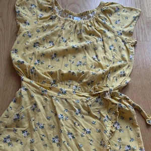 Gul blommig omlott-byxdress Lindex - Supersöt gul  omlott-byxdress från Lindex med små blommor i blått och vitt. Omlott-byxdress har omlottmodell med volangkant och knyt i midjan. Perfekt för sommarens alla dagar, med korta ärmar och luftig känsla. Tillverkad i mjuk bomull.