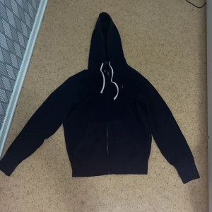 Mörkblå hoodie från Polo Ralph Lauren - Snygg mörkblå hoodie från Polo Ralph Lauren med vit snörning i huvan och röd broderad logga på bröstet. Tröjan har dragkedja framtill, känguruficka och är tillverkad i mjuk bomull. Perfekt för en chill och stilren look.