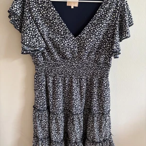 Blommig kortklänning - Supersöt kortklänning  med blå botten och vitt blommönster. Klänningen har v-ringning, volangärmar och smock i midjan för en skön passform. Perfekt för varma dagar och har en luftig känsla.