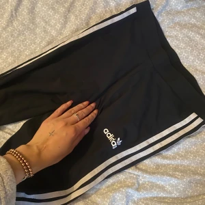 Svarta Adidas mjukisbyxor med ränder - Svarta mjukisbyxor från Adidas med klassiska vita ränder längs sidorna och Adidas-logga på benet. Byxorna har elastisk midja och är perfekta för en sportig och avslappnad stil.