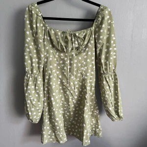 Grön blommig kortklänning med puffärm - Supersöt ljusgrön kortklänning med vita blommor, långa puffärmar och söt knytning vid bysten. Klänningen har en avslappnad passform och är perfekt för vår och sommar. Materialet känns lätt och luftigt, och den blommiga looken ger en romantisk vibe.