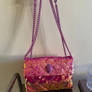 Paljettväska från Kurt Geiger - Färgstark axelväska från Kurt Geiger med glittriga paljetter i rosa, gult och orange. Väskan har en kedjerem i metallicrosa som går att bära kort eller långt och ett dekorativt spänne framtill. Insidan är fodrad i rosa material och loggan är präglad på insidan.