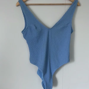 Blå ribbad baddräkt - Snygg blå baddräkt med ribbad struktur och djup v-ringning både fram och bak. Modellen har breda axelband och är tillverkad i ett stretchigt material som sitter skönt på kroppen. Perfekt för strandhäng eller poolparty.