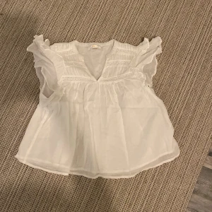 Vit blus med volang från Gina Tricot - Supersöt vit blus från Gina Tricot i luftig bomull. Blusen har volangdetaljer vid axlarna och v-ringning framtill. Perfekt för dig som gillar romantisk och feminin stil. Passar till både jeans och kjol!