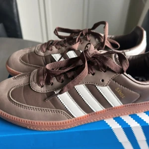 Adidas Samba bruna sneakers strl 40⅔ - Snygga Adidas Samba sneakers i brunt skinn och mocka med klassiska vita ränder och gummisula. Skorna har snörning och guldfärgad Samba-text på sidan. Hade behållt de själv om jag inte råkat beställa fel storlek 🥲