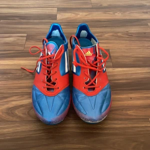 Adizero blå och röda fotbollsskor Messi 2012 - Snygga fotbollsskor från Adidas i blått och rött med silvriga ränder på sidorna. Skorna har snörning, platt sula med gröna dobbar och är tillverkade i syntetmaterial. Perfekta för dig som vill sticka ut på planen med en färgstark look.