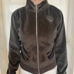 Svart velour bomberjacka med hjärtdetalj - Mjuk mörk grå bomberjacka i velour med dragkedja och fickor framtill. Jackan har en liten hjärtdetalj i strass på bröstet och ribbade muddar. Perfekt för dig som gillar en chill och snygg stil.