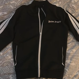 Palm Angels svart track jacket - Svart track jacket från Palm Angels med vita ränder längs ärmarna och vit dragkedja. Jackan har hög krage, två fickor med dragkedja och logga på bröstet. Helt utan problem, pris kan diskuteras 