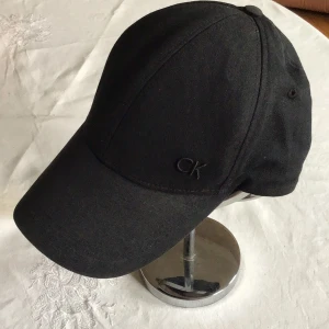 Svart keps från Calvin Klein - Stilren svart keps från Calvin Klein med diskret CK-brodyr framtill. Klassisk böjd skärm och justerbar metallspänne bak. Tillverkad i bomull för en bekväm känsla. Perfekt accessoar för en clean och modern look.passar för liten huvud nästan barn  storlek .