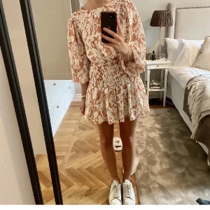 Beige och vit mönstrad kortklänning - (Köpt på Plick!!❤️)Säljer en söt kortklänning i beige och vitt med blommigt mönster. Klänningen har trekvartsärm, markerad midja med smock och volang i kjolen. Perfekt i sommar❤️