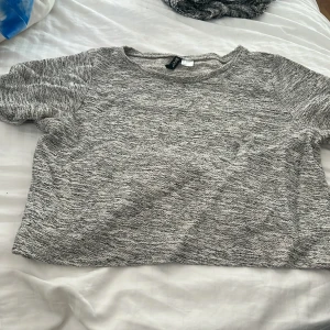 Grå croppad topp från Divided - En enkel och stilren grå croppad topp från Divided. T-shirten har korta ärmar och är tillverkad i ett mjukt, strukturerat material som känns skönt mot huden. 