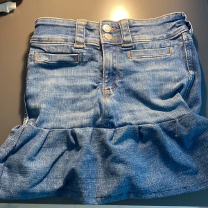 Blå jeanskjol med volangkant - Snygg blå jeanskjol med volang längst ner och dubbla knappar i midjan. Jag har sytt kjolen själv så den är inte helt perfekt sydd men inget man märker!