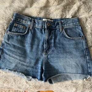 Blå jeansshorts från Pull&Bear - Klassiska blå jeansshorts från Pull&Bear med råa kanter och femficksmodell. Perfekta för sommardagar och enkla att matcha med allt. Materialet är slitstarkt denim i bomull.