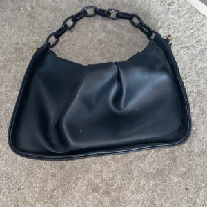 Svart axelväska med kedjehandtag - Säljer en svart axelväska i skinnimitation med chunky kedjehandtag i plast. Väskan har en snygg, slouchy form och stängs med dragkedja i guld. Perfekt storlek för det viktigaste och ger en cool vibe till din outfit.jag har ingen aning vart den kommer ifrån ! 🥹
