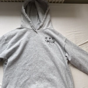Grå hoodie från Gina Tricot med tryck - Grå hoodie från Gina Tricot med svart texttryck 'JE M'EN AMOUR' och en stjärna både fram och bak. Mjuk insida, huva och långa ärmar. Perfekt för dig som gillar chill och enkel stil.