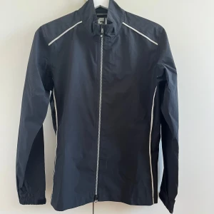Svart regnjacka från FootJoy - Svart regnjacka från FootJoy med vita detaljer längs axlar och sidor. Jackan har hel dragkedja framtill, hög krage och elastiska muddar vid ärmsluten. Perfekt för blåsiga dagar och sportiga outfits. Köpt för 1500 kr. 
