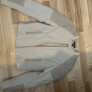 Beige jacka från H&M - Snygg beige jacka från H&M i storlek 38, men känns smal i ärmarna. Jackan har prydnads detaljer på axlar och ärmar, dragkedja framtill och två fickor. Perfekt för dig som gillar trendiga plagg.