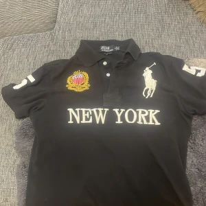 Svart pikétröja Polo Ralph Lauren S - Svart pikétröja från Polo Ralph Lauren i bomull, custom fit. Stora broderade detaljer med 'NEW YORK', emblem och polospelare framtill samt siffran 5 på ärmar och rygg. Klassisk krage och korta ärmar, perfekt för en sportig och stilren look.