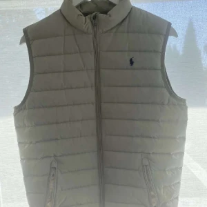 Beige dunväst från Polo Ralph Lauren  - Snygg grå dunväst från Polo Ralph Lauren i storlek XL juniorstorlek. Västen är quiltad med horisontella sömmar, har hög krage och dragkedja framtill. Klassisk blå logga broderad på bröstet och två fickor med dragkedja. Perfekt för lager-på-lager-stil. Skriv vid funderingar. 