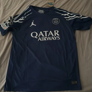 PSG matchtröja D. Doué #14 blå - Paris Saint-Germain matchtröja i mörkblått med vita detaljer, Jordan-logga och klubbmärke på bröstet. Kortärmad modell med tryckta sponsorloggor och Champions League-patch på ärmen. Namn och nummer D. Doué 14 på ryggen. Tillverkad i lätt och ventilerande material.