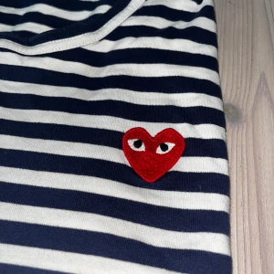 Randig tröja Comme des Garçons Play - Säljer en klassisk randig tröja från Comme des Garçons Play i marinblått och vitt. Tröjan har ett ikoniskt rött hjärta med ögon broderat på bröstet och är tillverkad i mjuk bomull. Perfekt för dig som gillar streetstyle och vill sticka ut.