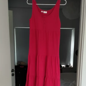 Rosa långklänning från bpc - Snygg långklänning i rosa från bpc. Klänningen har rund halsringning, knappar framtill och ett luftigt fall med flera volanger. Perfekt för varma dagar!
