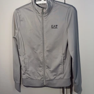 Grå track jacket från EA7 Emporio Armani - Snygg grå track jacket från EA7 Emporio Armani med svart logga på bröstet och stor vertikal logga på ryggen. Jackan har dragkedja hela vägen, ståkrage och två fickor framtill. Perfekt för en sportig och clean look.