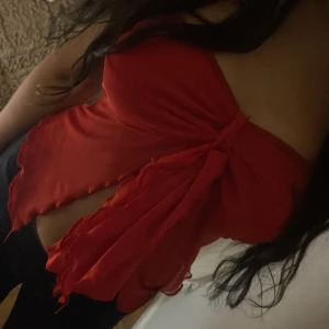 Röd bandeau tubtopp med volang - Säljer en röd bandeau tubtopp med volangdetaljer och knyt framtill. Toppen är ärmlös och har en flowy, luftig känsla som ger en snygg och trendig look. Perfekt för dig som vill sticka ut med en färgstark och unik topp.