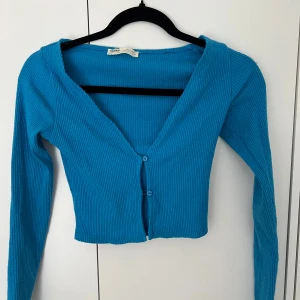 Blå ribbad kofta från Pull&Bear - Snygg blå ribbad kofta från Pull&Bear med croppad passform och v-ringning. Koftan har långa ärmar och stängs med två knappar framtill. Perfekt att styla med jeans eller kjol för en chill look.