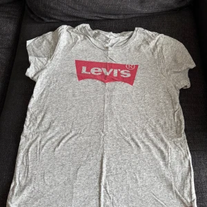 Grå Levi's t-shirt med rött tryck - Snygg grå t-shirt från Levi's med klassiskt rött logotryck på bröstet. T-shirten har rund hals, korta ärmar och är gjord i mjuk bomull. Perfekt för en chill och avslappnad stil.