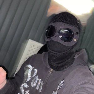 Svart balaclava med goggles C.P. Company - Unik svart balaclava från C.P. Company med inbyggda svarta goggles och stickad design. Huvan täcker hela huvudet och har en cool, futuristisk vibe. Perfekt för dig som vill sticka ut och hålla dig varm med stil. No size