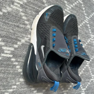 Nike Air Max 270 svart/blå sneakers - Nike Air Max 270 sneakers i svart med blå detaljer och vit sula. Ovandelen är i mesh för bra andningsförmåga och har elastiska band istället för klassisk snörning. Den ikoniska Air Max-sulan ger extra komfort och en sportig look. Perfekta för dig som gillar streetstyle.