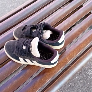 Svarta Adidas Campus sneakers - Klassiska Adidas Campus sneakers i svart mocka med vita ränder och vit sula. Skorna har snörning och rund tå, samt Adidas-logga på plösen och hälen. Perfekta för en avslappnad streetstyle-look.