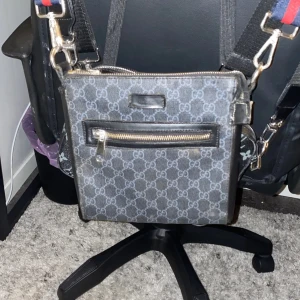 Grå axelväska med mönster - Säljer en snygg grå axelväska med ett stilrent mönster. Väskan har en praktisk dragkedja framtill och en justerbar axelrem i blått och rött. Perfekt för både vardag och fest! Det är lite tröga dragkedjor men annars inga konstigheter!