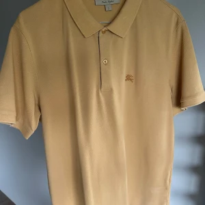 Beige Burberry pikétröja i bomull - Snygg beige pikétröja från Burberry i 100% bomull. Klassisk krage, korta ärmar och knäppning med Burberrys ikoniska rutiga detalj på insidan. Liten broderad logga på bröstet. Perfekt för en clean och stilren look.