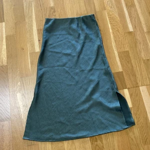 Mörkgrön långkjol med slits - Snygg mörkgrön långkjol i slät, glansig kvalitet med en stilren slits på sidan. Kjolen har en rak passform och faller snyggt längs benen. Perfekt för dig som gillar minimalistisk stil och vill ha något enkelt men ändå trendigt.