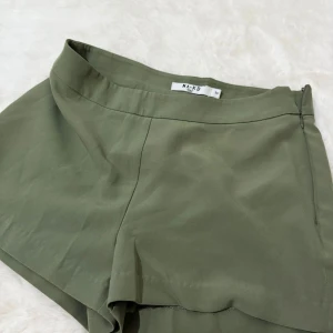 Olivgröna shorts från NA-KD - Stilrena olivgröna shorts från NA-KD med hög midja och diskret dragkedja i sidan. De har en enkel, clean look och är tillverkade i ett mjukt, lätt material som känns skönt mot huden. Perfekta för varma dagar när du vill ha en chill men snygg stil.