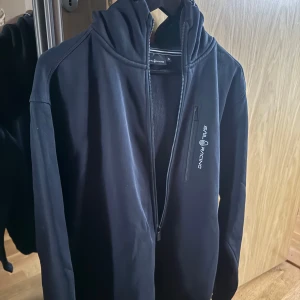 Svart vindjacka från Sail Racing XL - Snygg grå hoodie från Sail Racing i storlek XL. Tröjan har dragkedja, huva och en bröstficka med zip. Insidan är mjukt fodrad med fleece som gör den mycket skön. Perfekt till casual tillfällen eller bara att ha hemma. Den är nästintill aldrig använd och är i mycket bra skick. Ordinarie pris 1100.