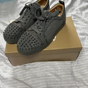 Gråa Christian Louboutin sneakers med nitar - Snygga grå sneakers från Christian Louboutin med coola svarta nitar på tån och klassisk röd sula med logga. Skorna har snörning och är tillverkade i mocka för en lyxig känsla. Perfekta för dig som vill sticka ut med en edgy stil. Kvitto finns. Köpte i Köpenhamn 