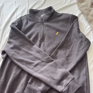 Grå half zip sweatshirt från Lyle & Scott - Mörkgrå sweatshirt från Lyle & Scott med halv dragkedja och broderad gul logga på bröstet. Tröjan har lång ärm, hög krage och är gjord i mjuk bomull. Perfekt för en chill och stilren look.