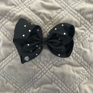 Jojo Siwa -Svart diadem med rosett och pärlor - Stort svart diadem med en maffig rosett dekorerad med glittrande pärlor. Rosetten har en snygg struktur och en liten silverfärgad detalj i ena hörnet. Perfekt accessoar för att lyfta din look.