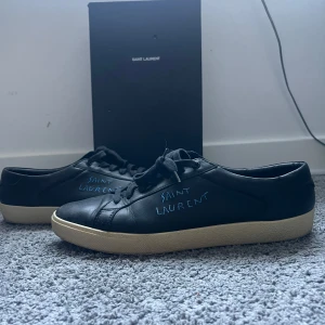  Saint Laurent  - Svarta sneakers från Saint Laurent i mjukt skinn med handskriven logga i blått på sidan. Klassisk låg modell med svarta snören och vit platt sula. Snygg minimalistisk design som passar till allt. Levereras med originalkartong och dustbags. Och extra skosnören följer med och so. Är i storleken 46