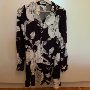Svartvit blommig skjortklänning H&M - Svart skjortklänning från H&M med stora vita blommor och blad. Klänningen har knappar framtill, lång ärm med manschett och en lös, flowy passform. Helt oanvänd och köpt från årets kollektion.