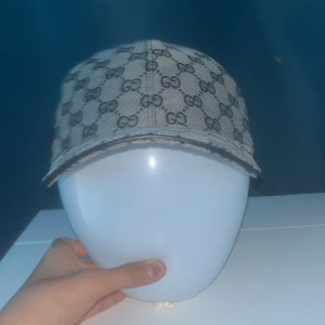 Gucci keps med GG-mönster och röd rand - Snygg keps från Gucci i beige och mörkblått med klassiskt GG-monogram över hela kepsen. På sidan finns en ikonisk röd och marinblå rand. Justerbar rem baktill i skinn och insida i mörkblått tyg. Perfekt för dig som vill sticka ut med lyxig streetstyle.