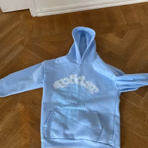 Ljusblå hoodie med tryck och huva - Säljer en ljusblå hoodie med coolt vitt tryck framtill som ser ut som bubbliga bokstäver och ett spindelnätsmönster. Tröjan har huva och en stor magficka. Perfekt för dig som gillar streetwear och vill sticka ut lite extra.