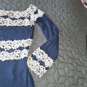 Marinblå spetsklänning från Intimissimi - Snygg marinblå kortklänning från Intimissimi med långa ärmar och offshoulder-modell. Klänningen har breda band av vit blommig spets över bröst, ärmslut och midja. Mjuk och stretchig bomullsblandning, perfekt för dig som gillar romantiska detaljer. Den är I mörk blå men på bilden ser ljusare ut på grund av kameran. 