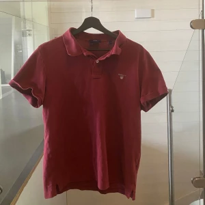 Röd pikétröja från GANT - Säljer en klassisk röd pikétröja från GANT med broderad logga på bröstet. Tröjan har krage, korta ärmar och knäppning framtill. Perfekt för en stilren och avslappnad look. Tillverkad i mjuk bomull för skön komfort.